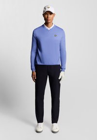 Maglione azzurro chiaro a V, camicia bianca con colletto, pantaloni scuri, scarpe bianche e guanto. Accento giallo del logo sul maglione e sul cappello.