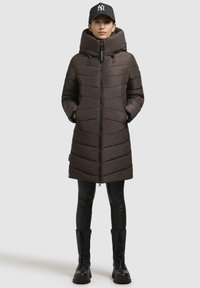 Cappotto lungo marrone imbottito con cappuccio, caratterizzato da una texture a quadri orizzontali, zip frontale e due tasche laterali. Stivaletti da combattimento neri alte.