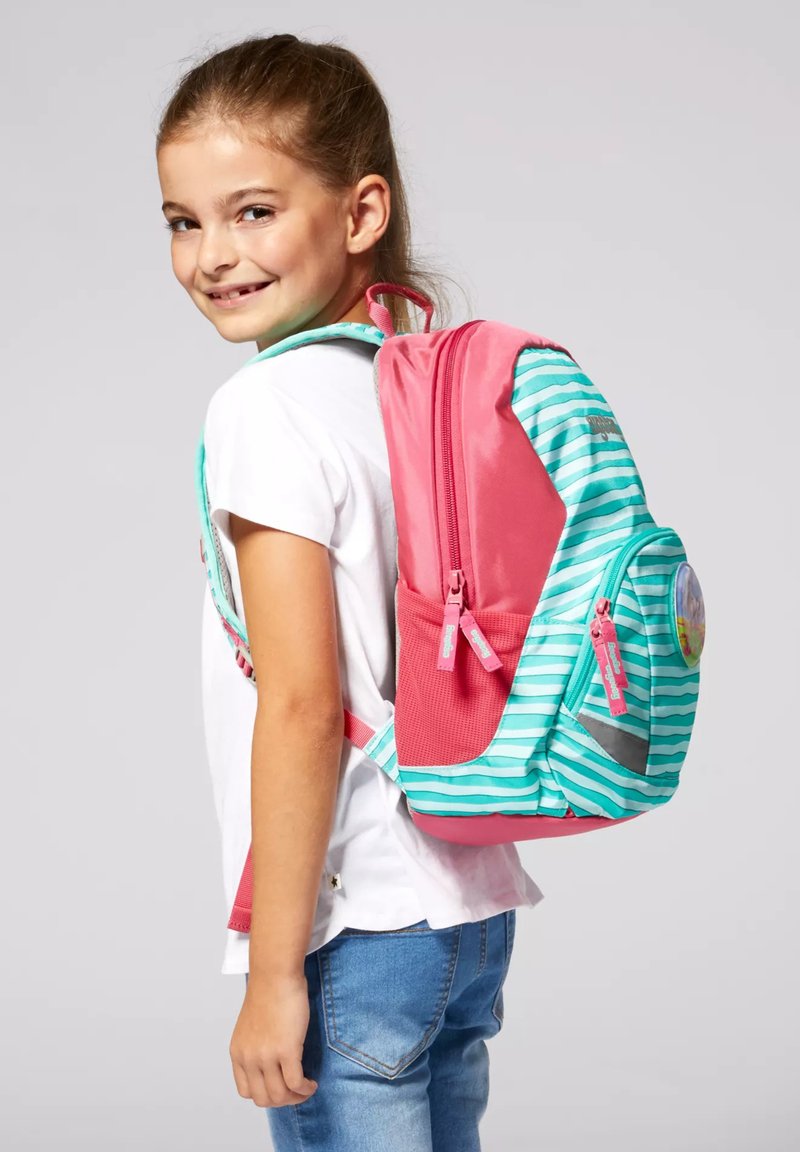 Lächendes Mädchen mit weißem Hemd und blauen Jeans trägt rosa-türkis gestreiften Rucksack mit mehreren Fächern.