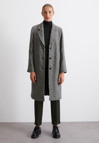 Palton lung alb-negru cu model houndstooth, cu rever notate, buzunare frontale și croială lejeră, asortat cu un guler înalt negru și pantaloni subțiri.