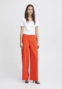 Witte aansluitende top met korte mouwen, gecombineerd met hooggebloemde, wijde oranje broeken. Glad materiaal, minimalistisch ontwerp en neutrale zwarte hakken.