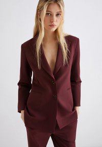 Giovane donna con lungo capelli biondi, che indossa un blazer scuro bordeaux su misura e pantaloni abbinati, in piedi davanti a uno sfondo chiaro e uniforme.