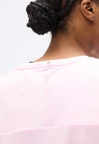 Αθλητική μπλούζα ανοιχτό ροζ με υφή σε σχήμα βέλους, με ένα μικρό λογότυπο στο πίσω μέρος του λαιμού. Μαλακό ύφασμα με λεία επιφάνεια.