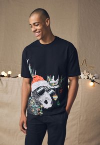 Black t-shirt featuring a graphic of a moose with sunglasses, wearing a red Santa hat, adorned with festive decorations like ornaments and lights.