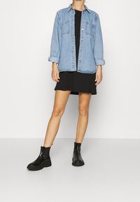 Femme portant une robe noire, une veste en denim bleu clair à manches retroussées et des bottines noires, adossée à un fond clair.
