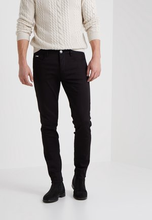 Hombre con pantalones ajustados negros, suéter de punto trenzado color crema y botas negras hasta el tobillo, de pie contra un fondo blanco liso.