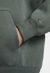 Sweatshirt verde escuro com tecido texturizado, apresentando um bolso e um pequeno logótipo bordado em preto na manga. Design suave e casual.