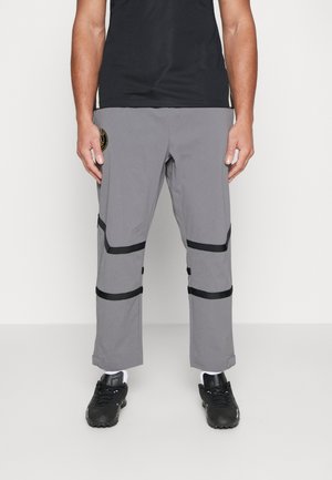 Osoba w szarych spodniach z czarnymi paskami powyżej kolan i łydek, czarne trampki z białymi skarpetkami, stojąca na jednolitym tle.