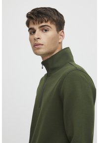 Grön zip-up hoodie gjort av texturerat tyg med hög krage. Har en halvvista stängning och synliga sömdetaljer.