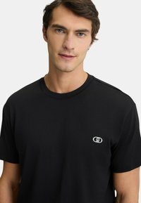 T-shirt nera a maniche corte in cotone, con un piccolo logo bianco nella zona sinistra del petto e un classico scollo rotondo.