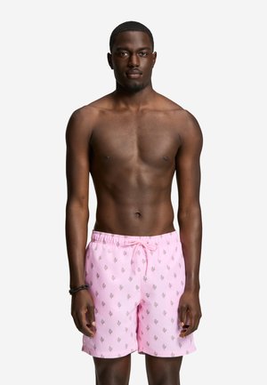 Uomo senza maglietta che indossa pantaloncini da bagno rosa con piccoli motivi vegetali scuri, in piedi di fronte a uno sfondo chiaro e uniforme.