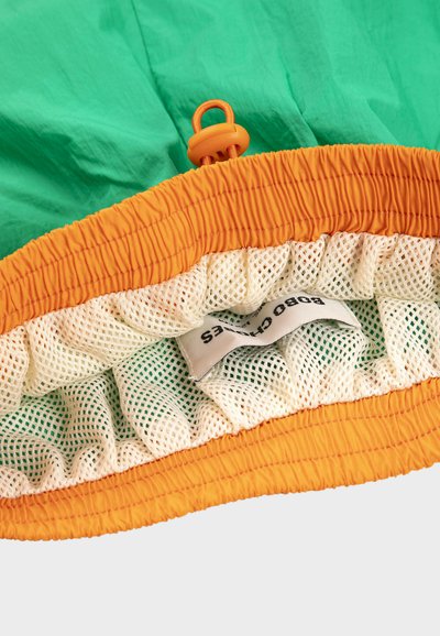 Gros plan sur une ceinture élastique orange avec un cordon de serrage réglable, une doublure en filet blanc et une étiquette de vêtement partiellement visible à l'intérieur d'un tissu vert.
