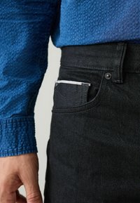 Svarta denimbyxor med smal passform, utrustade med en tydlig vit blixtlåsficka och en delvis synlig texturerad blå skjorta.