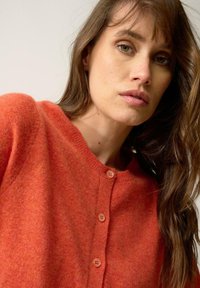 Orange stickad kofta med rund halsringning, utrustad med tre knappstängningar och en mjuk textur. Detaljer inkluderar subtila färgvariationer.