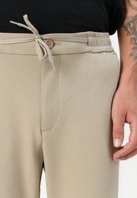 Beige housut, joissa on joustava vyötärö ja kiristysnauha, sileällä pinnalla ja sivutaskuilla, näytetty vyötärölle asti.