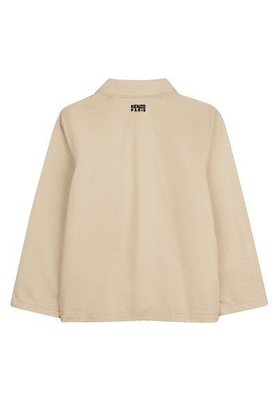 Beige jas met een brede kraag en lange mouwen. Heeft een zwart logo op de bovenrug. Gladde textuur en een losse pasvorm.