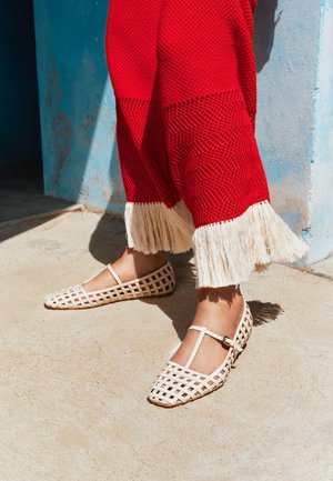 Πόδια με λευκά παπούτσια Mary Jane με σχέδιο πλέγματος και κόκκινο υφασμάτινο παντελόνι με λευκή φούντα, που στέκονται σε ηλιοφώτιστο τσιμεντένιο δάπεδο.