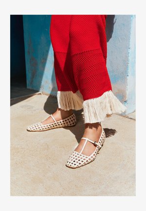 Stopy w białych butach Mary Jane z wzorem kratki i czerwonych teksturowanych spodniach z białym frędzlem, stojące na oświetlonej słońcem betonowej podłodze.