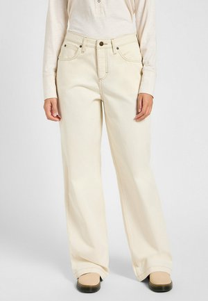 Blusa de punto en crema de manga larga con tapeta de botones, combinada con pantalones beige de pierna ancha y cintura alta. Sosteniendo una camisa de franela a cuadros en tonos rosa y azul.