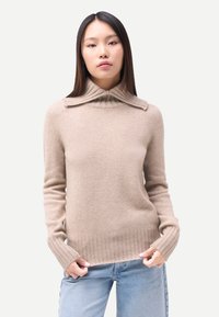 Femme debout portant un pull à col roulé côtelé beige et un jean bleu clair, sur un fond blanc uni.