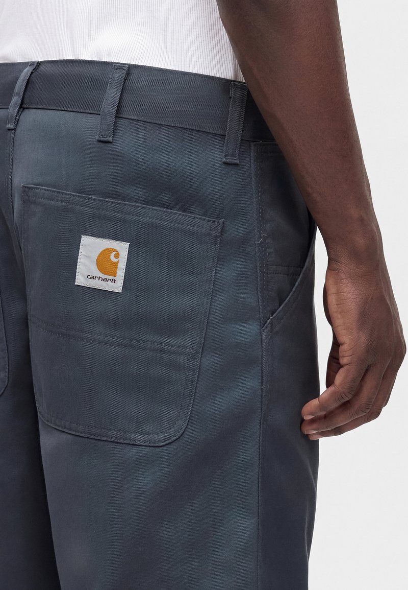 Gros plan d'une personne portant un pantalon Carhartt gris foncé avec une poche arrière carrée et un patch logo de la marque visible.