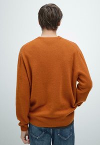 Brązowy sweter z dzianiny o luźnym kroju, z prążkowanymi mankietami i dolnym brzegiem. Tył ma zaokrąglony dekolt i miękką teksturę, noszony z dżinsami.