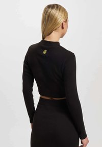 Svart kortermet topp med lange ermer laget av ribbestrikket materiale med en tettsittende design. Har en liten gul logo på baksiden av halslinningen.