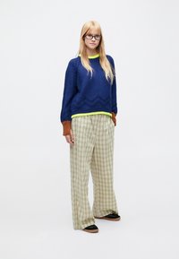 Maglione lavorato a maglia blu navy con motivo a zigzag e rifinitura verde neon; pantaloni a gamba larga a quadri verdi e crema; scarpe nere con suole color cuoio.