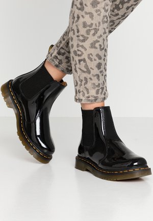 Personne portant des bottines Chelsea noires brillantes et un pantalon gris à imprimé léopard, debout sur un sol blanc lisse.