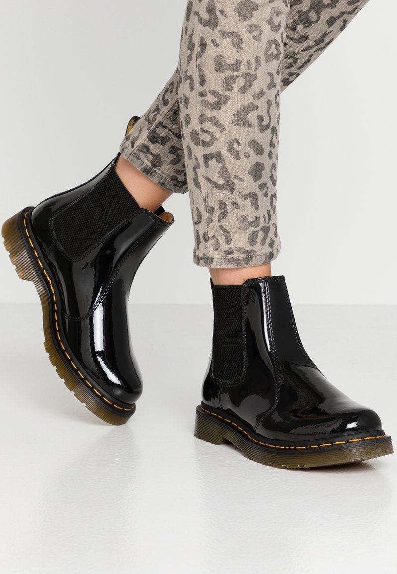 Personne portant des bottines Chelsea noires brillantes et un pantalon gris à imprimé léopard, debout sur un sol blanc lisse.
