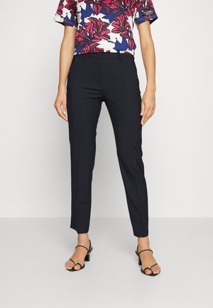 Osoba nosząca dopasowane czarne spodnie do kostki, kwiecistą koszulę z krótkim rękawem z czerwonymi i niebieskimi wzorami oraz czarne sandały na obcasie z paskami.