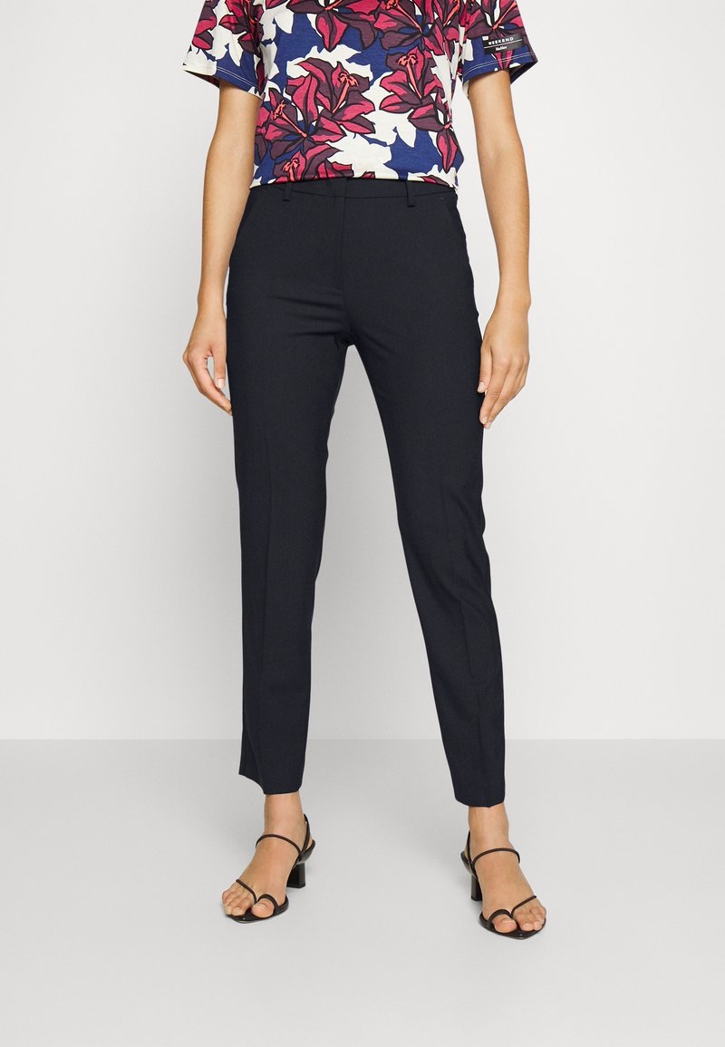 Personne portant un pantalon noir ajusté à la cheville, une chemise à manches courtes à motifs floraux rouges et bleus, et des sandales noires à talons avec des lanières.