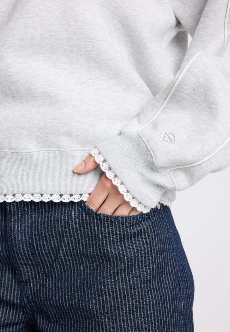 Sweat-shirt gris avec une bordure en dentelle aux ourlets et aux poignets, associé à un denim rayé foncé. Présente une texture côtelée et un détail logo.