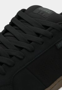 Μαύρο αθλητικό παπούτσι με κλωστοϋφαντουργικό άνω μέρος από υφή μαύρου υφάσματος, επίπεδες μαύρες κορδόνες και καφέ καουτσούκ σόλα με λεπτομέρειες κοπής και ετικέτα λογότυπου.