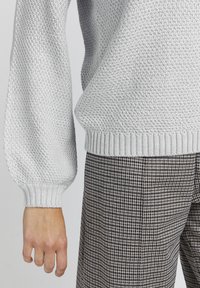 Hellgrauer, gestrickter Pullover mit strukturiertem Muster, mit gerippten Bündchen und Saum. Kombiniert mit braunen Karohosen mit kleinen Checks.