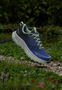 Sinine ja must Saucony jooksujalats kergete roheliste paeltega, hõljuv kivisel rajal, mida ümbritseb roheline taimestik.