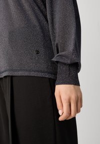 Nahaufnahme einer Hand einer Person an ihrer Seite, die ein dunkel schimmerndes Langarmoberteil mit gerippten Bündchen und schwarze strukturierte Hosen trägt.