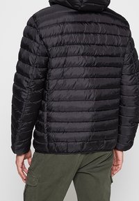 Sort pufferjakke med hætte, med horisontal quilting, blank overflade og elastiske manchetter. Båret over grønne bukser.