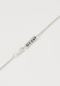 Bracelet en chaîne argentée avec une étiquette rectangulaire présentant des lettres noires en relief épelant "ICON". Le fermoir est de type homard.