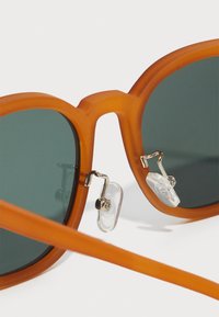 Gafas de sol naranjas mate con lentes verdes, que presentan un diseño redondeado ligeramente sobredimensionado y almohadillas nasales plateadas para mayor comodidad.