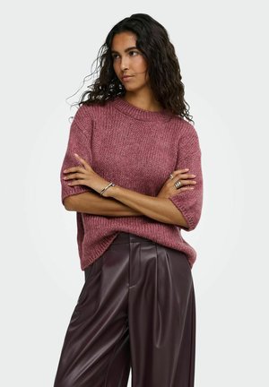Femme aux longs cheveux bouclés portant un pull en maille court mauve et un pantalon en cuir violet foncé, bras croisés, regardant sur le côté.