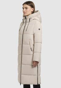 Heller beige langer gepolsterter Mantel mit Kapuze, ausgestattet mit einem Frontreißverschluss, verstellbaren Kordelzügen und Seitentaschen; glatte Textur und gestepptes Design.