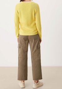 Gelb gestrickter Pullover mit langen Ärmeln, geripptem Muster, kombiniert mit hellbraunen, weiten Cargo-Hosen mit Seitentaschen. Sichtbares lässiges Schuhwerk.