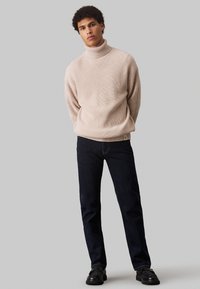 Pull en maille côtelée beige à col montant avec une coupe décontractée, associé à un jean droit bleu marine et des chaussures noires. Design simple et minimaliste.