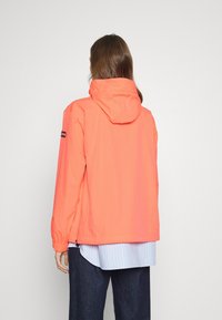 Korallfarbene Windjacke mit Kapuze, langen Ärmeln, elastischen Bündchen und lockerer Passform. Trägt man über einem hellblauen, gestreiften Hemd.