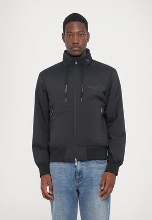 Mann iført en svart jakke med glidelås og snor, med frontlommer, sammen med lyseblå jeans, som står foran en hvit bakgrunn.