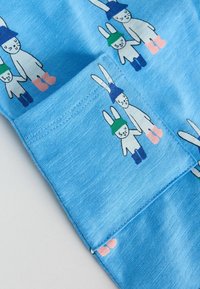 Tissu bleu avec une poche cousue, orné de paires de lapins blancs portant des chapeaux bleus et verts et des bottes colorées.