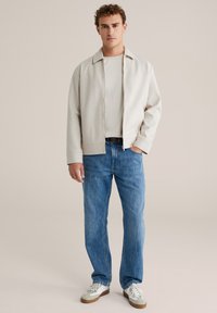 Chaqueta gris claro con cremallera, camiseta beige, vaqueros azul de pierna recta y zapatillas blancas con detalles grises. Tela suave y ajuste holgado.