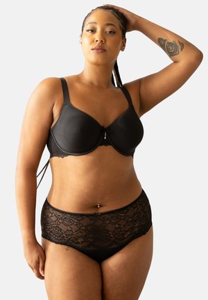 Frau mit geflochtenem Haar, die schwarzen BH und Spitzenunterwäsche trägt, posiert mit einem erhobenen Arm und zeigt ein Arm-Tattoo.