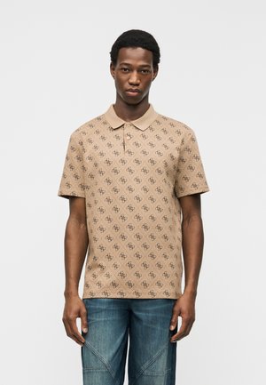 Άνδρας που φοράει μπεζ πουκάμισο πόλο με σχέδια και μπλε τζιν, στέκεται μπροστά σε απλό ανοιχτόχρωμο φόντο, κοιτάζοντας προς τα εμπρός με ουδέτερη έκφραση.